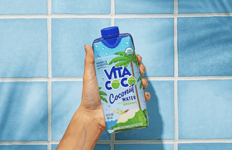Image 1: Une main présente un carton d'eau de coco Vita Coco sur un fond de carrelage bleu, soulignant son attrait dans le monde compétitif du marketing des boissons CPG.